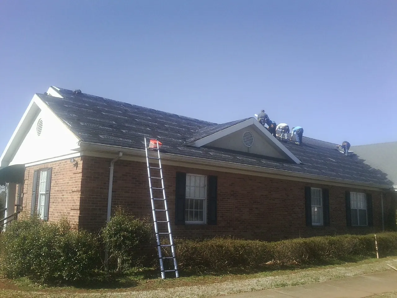 Crew tearing shingles off a brick ranch roof with a ladder set — Emergency Roof Repair work by our crew in Desmet