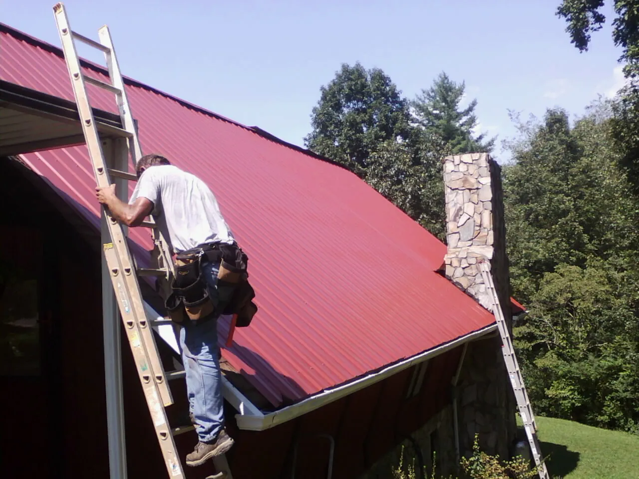 Roofer on a ladder beside a red metal roof and stone chimney — Soffit & Fascia Repair by Roofing Brothers in Desmet